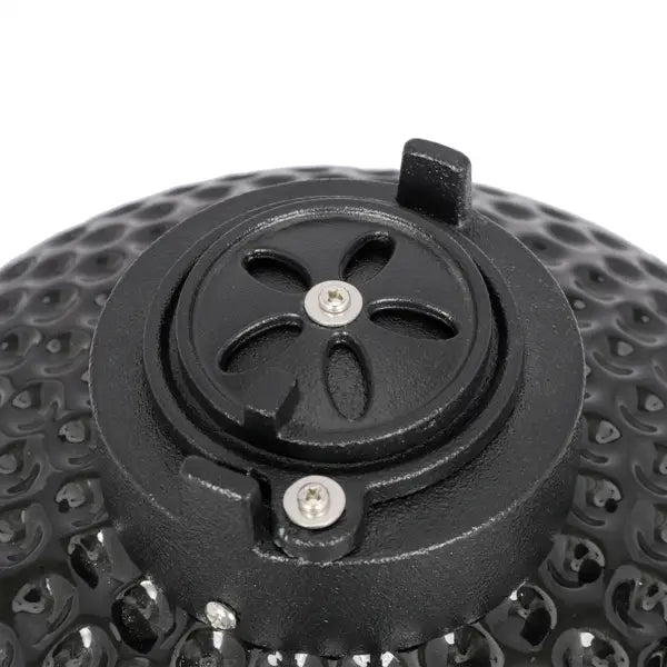 Black cast iron lid of a 13 inch ceramic kamado charcoal grill featuring a textured honeycomb pattern and a central flower-shaped vent cover secured by two silver screws.
