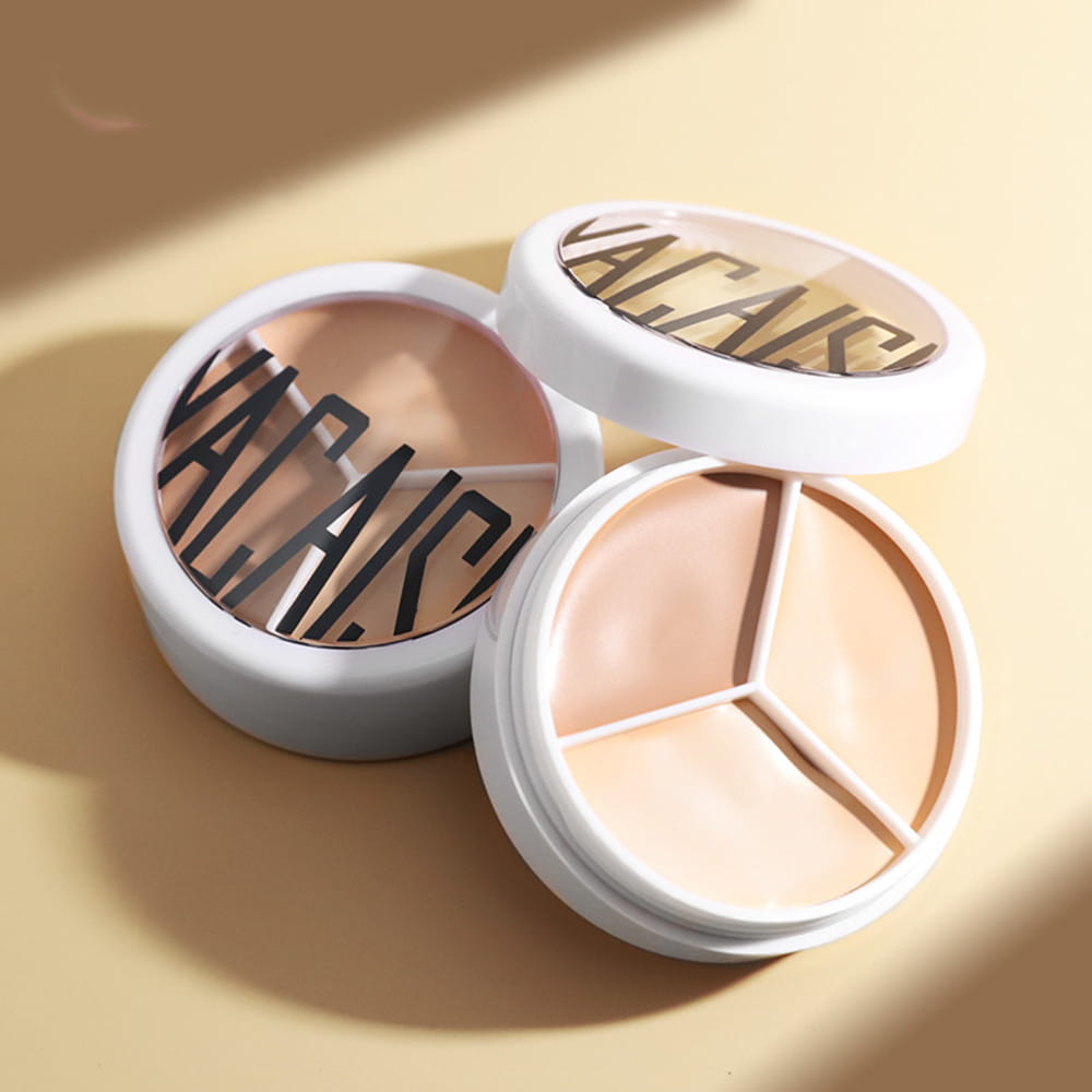 White circular three-color concealer palette with three-toned beige shades separated by a geometric design and branded Vacai on the lid, accompanied by a Pro 270 brush.