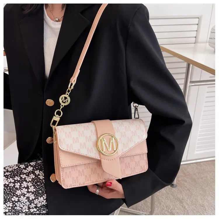 A blush pink monogram letter print crossbody messenger bag with gold-tone hardware and an adjustable strap featuring matching circular charms.