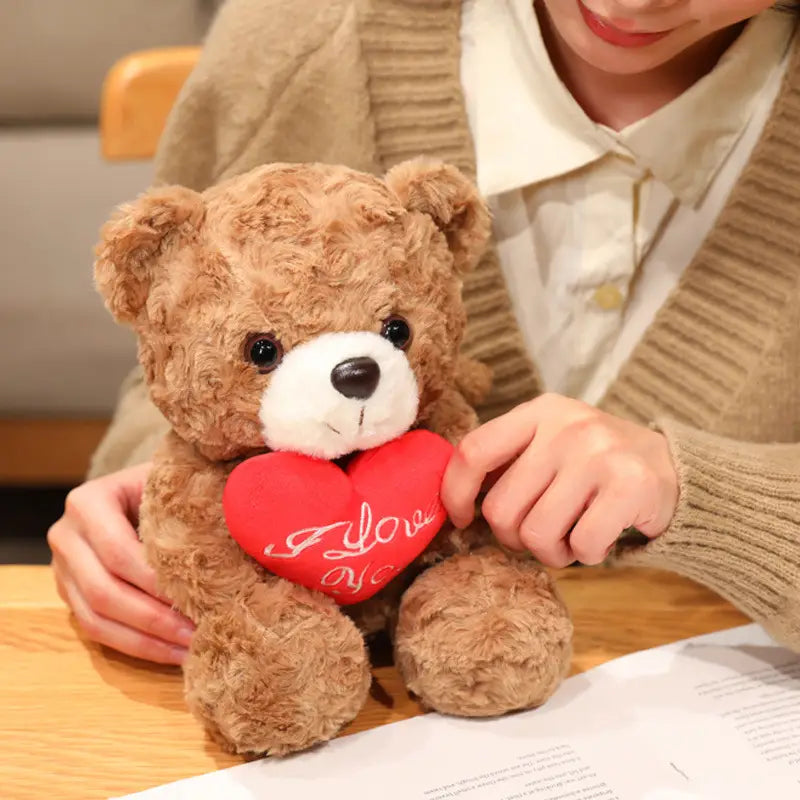 Ein brauner Plüsch-Teddybär mit weißer Schnauze und schwarzen Augen hält ein rotes Herz mit dem Text „I love you“ in Schreibschrift; präsentiert als Teddybär-Plüschtier mit Rosen, Valentinsgeschenk, 16–25 cm.