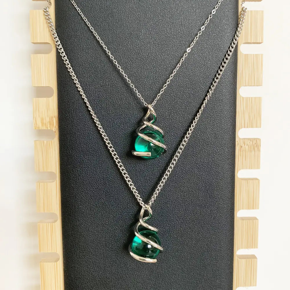 Dos collares de cadena de plata, cada uno con un colgante esférico de vidrio verde esmeralda vibrante, encapsulado en un diseño intrincado de espiral de alambre de plata retorcido.