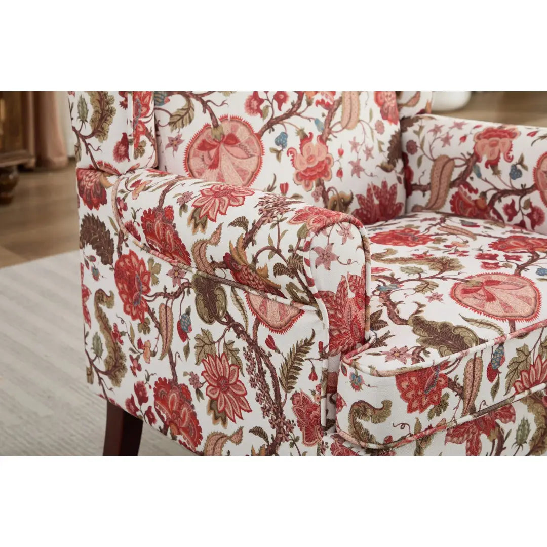 Una silla tipo ala con respaldo capitoné floral, tapizada en un tejido vibrante con flores rosadas rojas y marrones llamativas y hojas verdes sobre un fondo blanco, con patas de madera retro.
