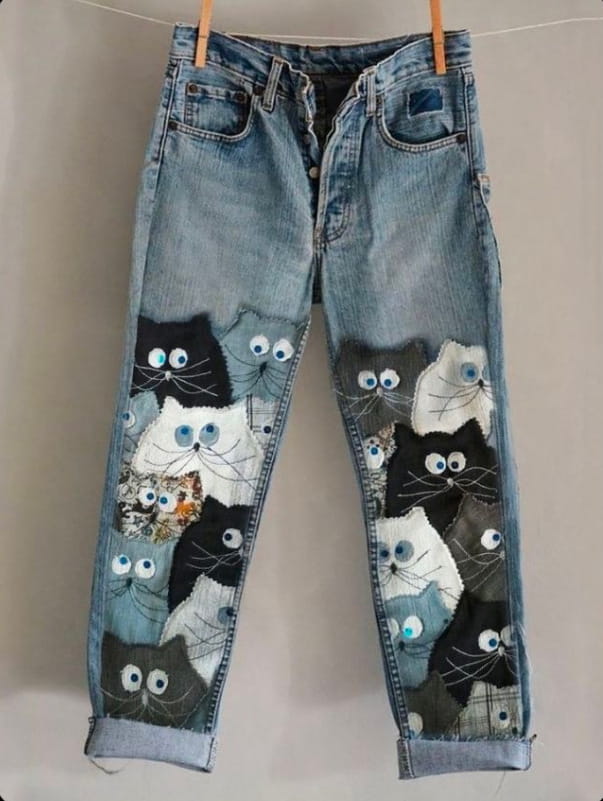 blue denim flared trousers featuring a patchwork of embroidered black white and gray cat faces on faux denim material
