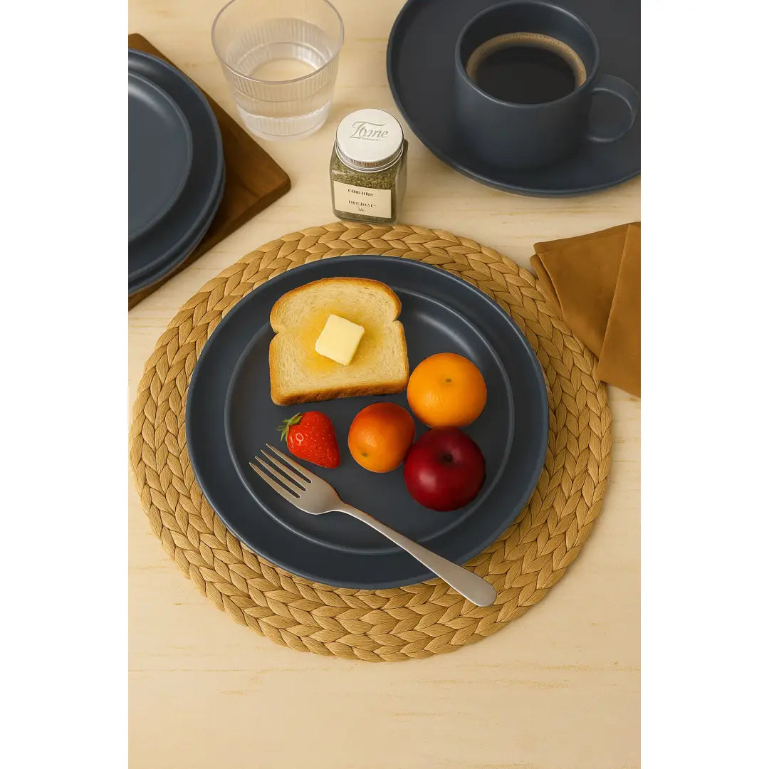 Plato de cerámica de loza azul oscuro con acabado mate que sostiene tostadas con mantequilla y frutas variadas de un juego de vajilla gris mate de 12 piezas para cuatro personas.