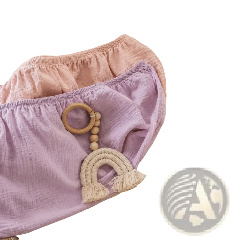 A breathable fitted baby crib mattress cover in soft lavender muslin with a wooden rainbow teether attached