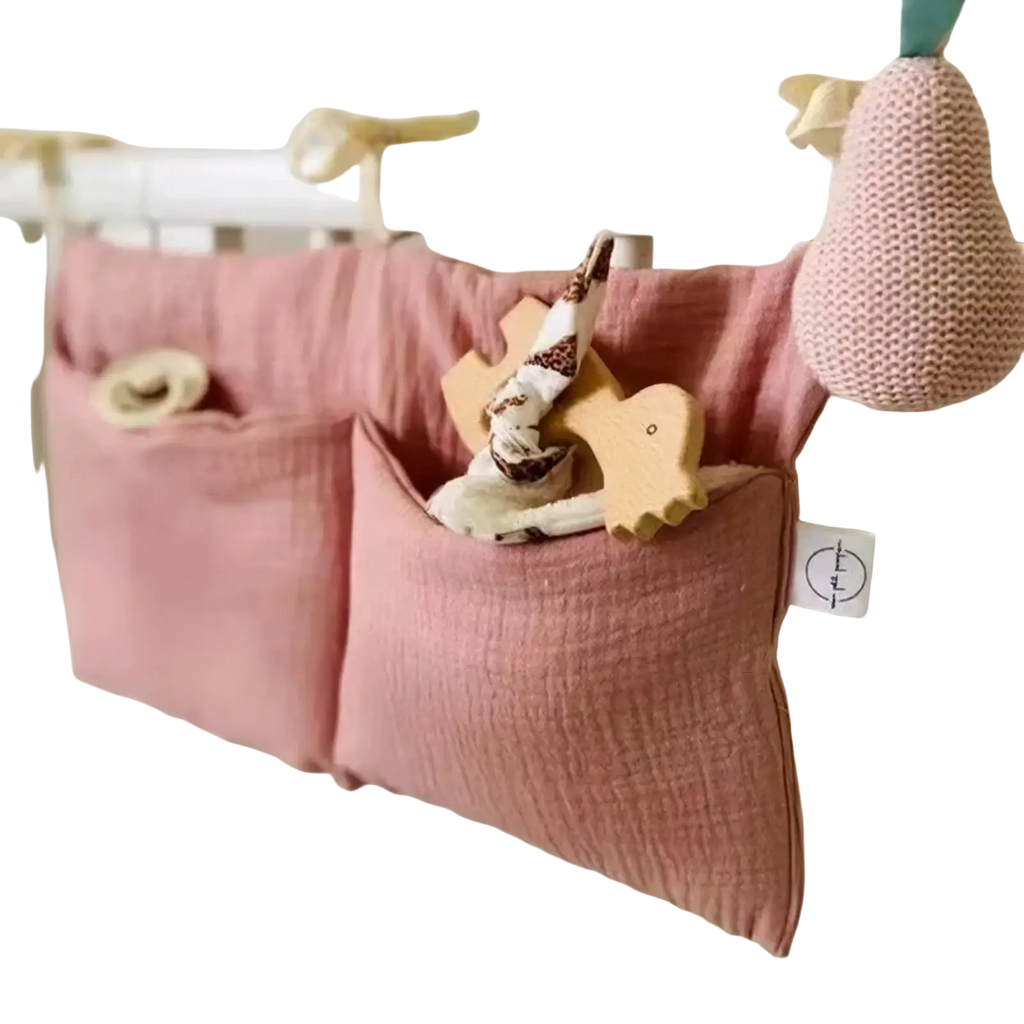 Pink cotton baby crib storage bag with two pockets hanging on white crib rail for bedside organization