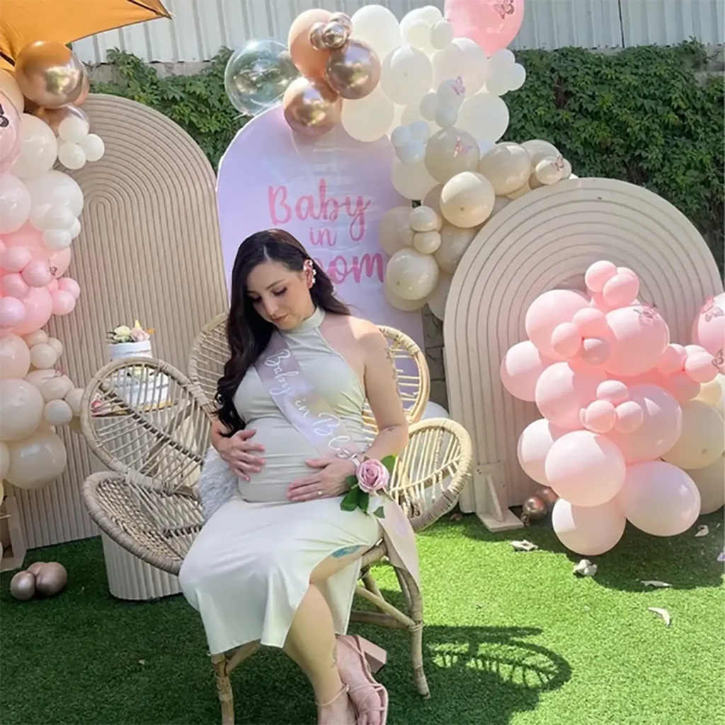 A pregnant woman wearing a blush pink satin baby shower sash seated in a wicker chair with hands on belly