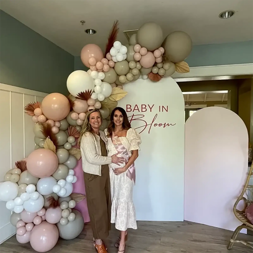 A baby shower sash in blush pink satin with a soft blush and cream balloon arch accented by dried pampas grass