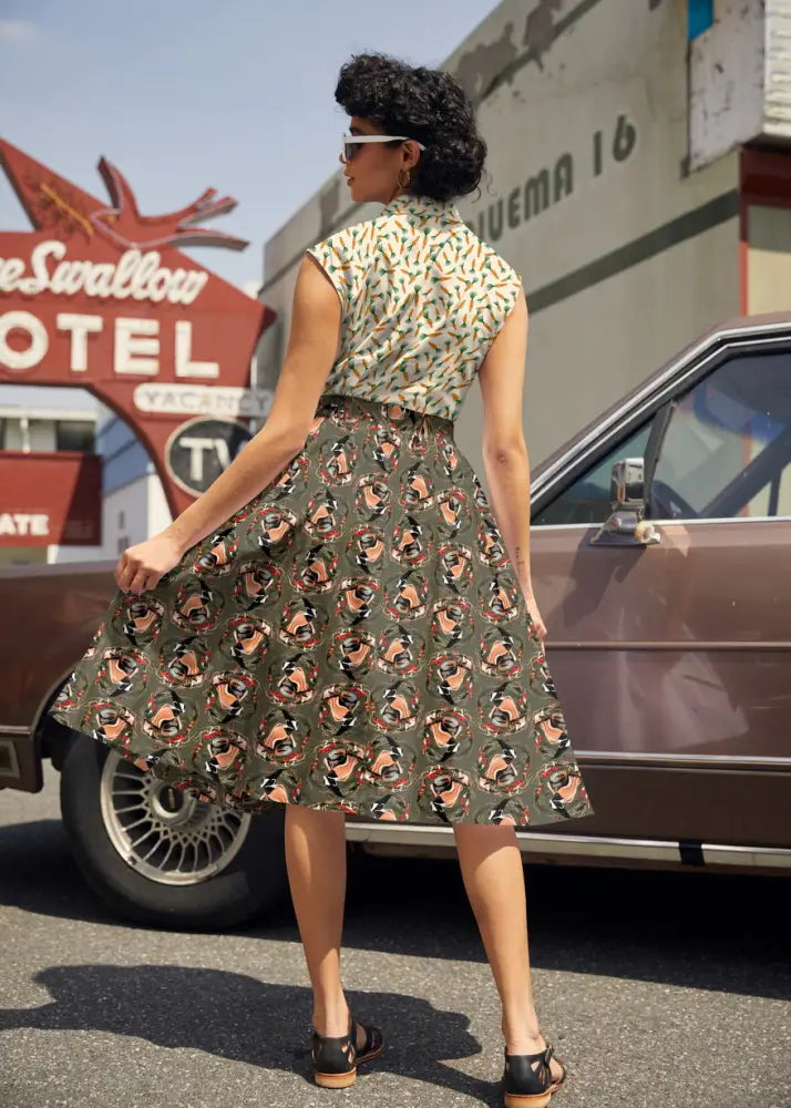 A woman wears a vintage-style sleeveless dress with green and beige pin-up face patterns and a Badger Doris olive green pleated cotton skirt with pockets