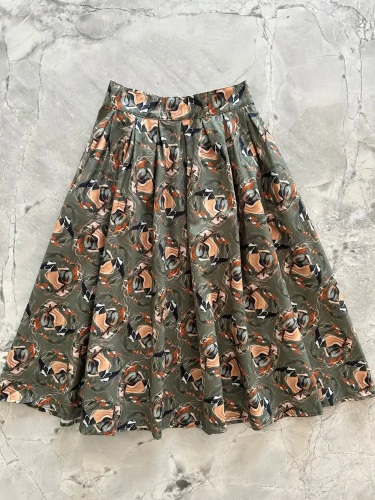 Badger Doris olive green pleated cotton skirt with pockets featuring a repeating pattern of abstract faces