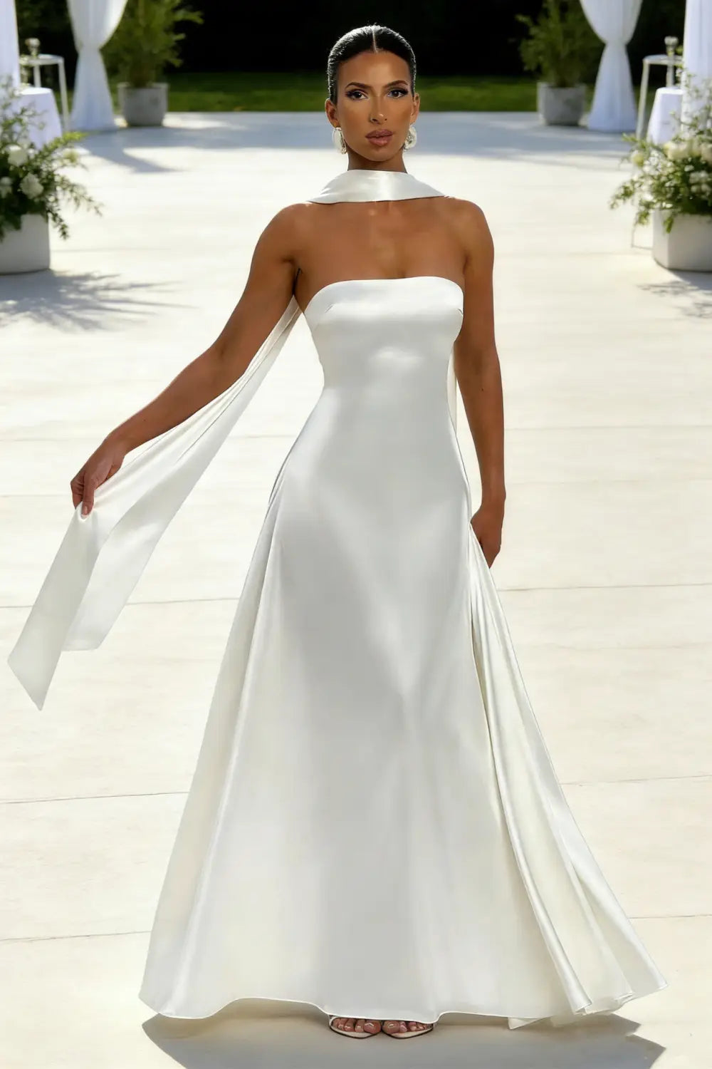 Un vestido de novia de satén blanco sin tirantes, de líneas elegantes, con un detalle tipo capa fluida en el hombro y un escote drapeado.