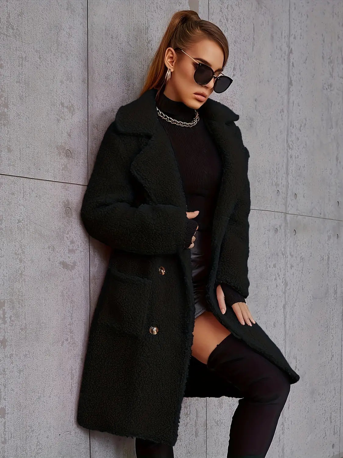 Manteau teddy mi-long doublé en polaire noire, avec un matériau en peau lainée, des boutons à ton or, et un col haut pour plus de chaleur et d’élégance.