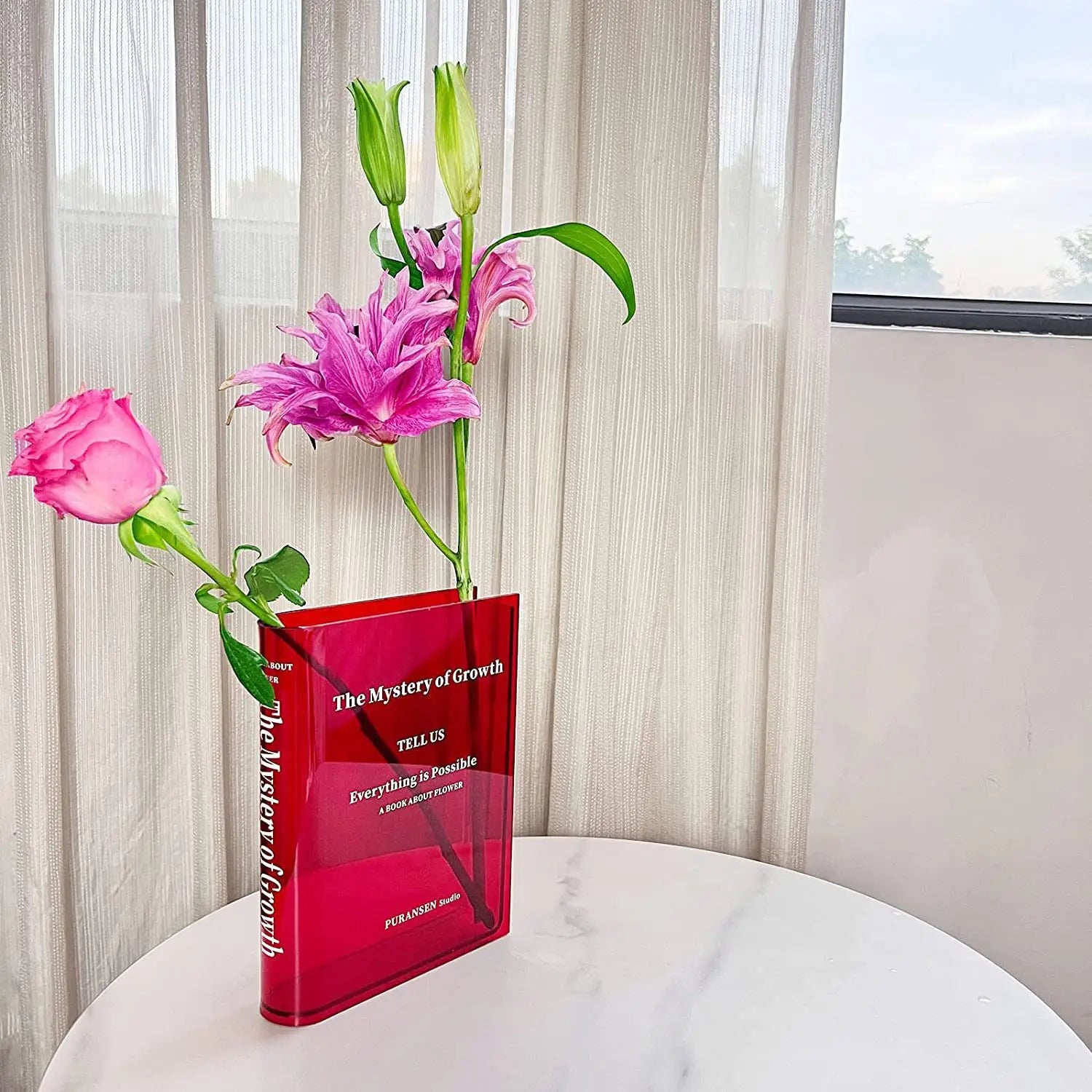 un jarrón de acrílico rojo translúcido con forma de libro y texto blanco que contiene flores rosadas y verdes para decoración moderna del hogar