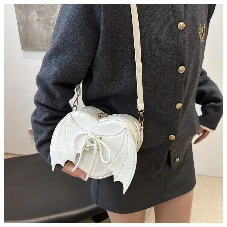 White bat wing heart crossbody bag with lace up detail and cream colored strap made of smooth material.
