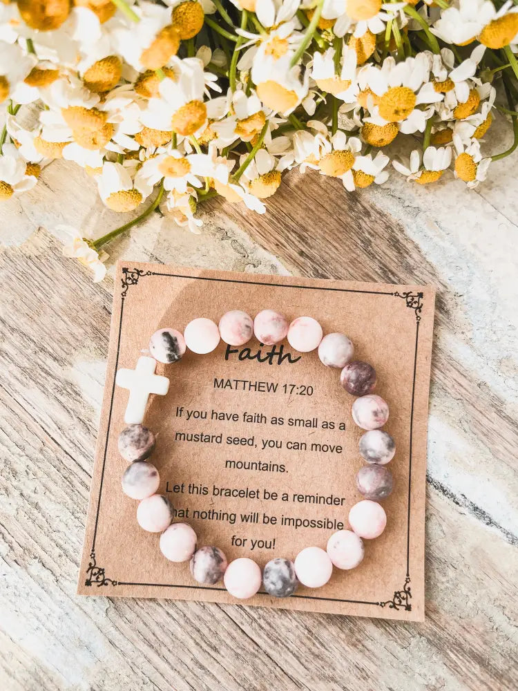 A pink and gray marbled stone stacking bracelet with a white cross charm on a rustic card with inspirational text