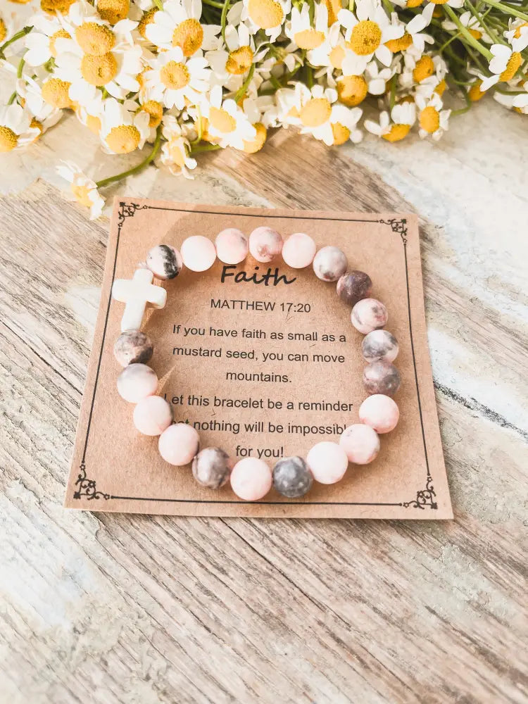A pink and gray marbled stone stacking bracelet with a white cross charm on a rustic display card