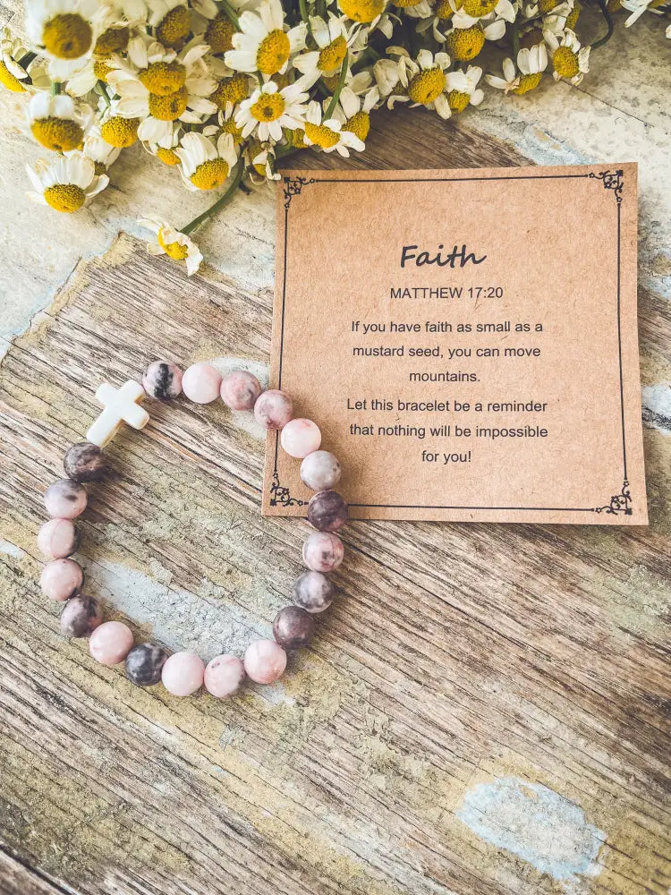 A pink and gray marbled stone stacking bracelet with a white cross charm resting beside an inspirational card