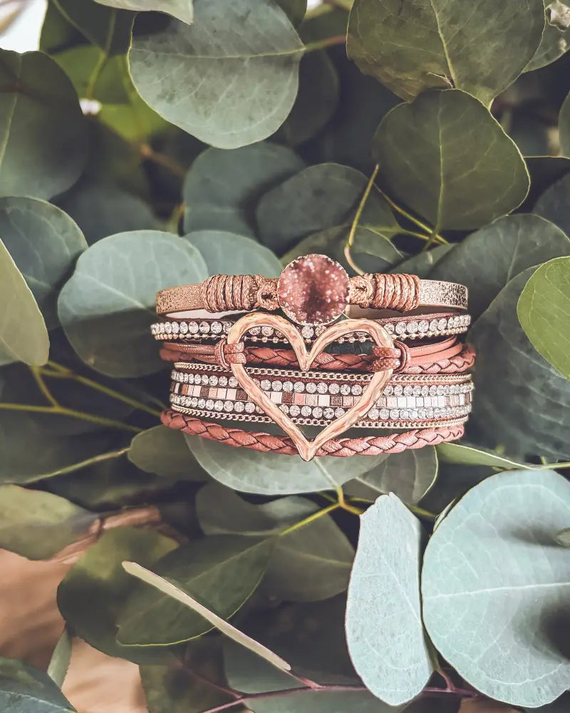 A stack of rose gold and copper-toned braided leather bracelets with a heart charm featuring clear crystals