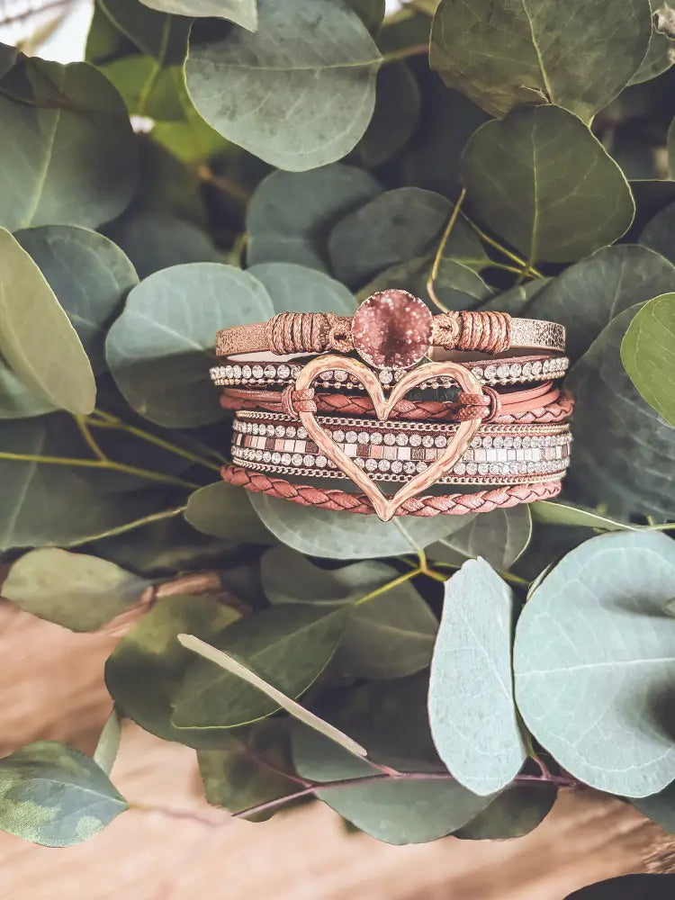 A stack of rose gold braided leather bracelets with a central heart charm featuring embedded crystals