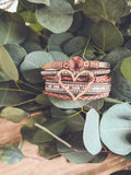 A stack of rose gold braided leather bracelets with a central heart charm featuring embedded crystals