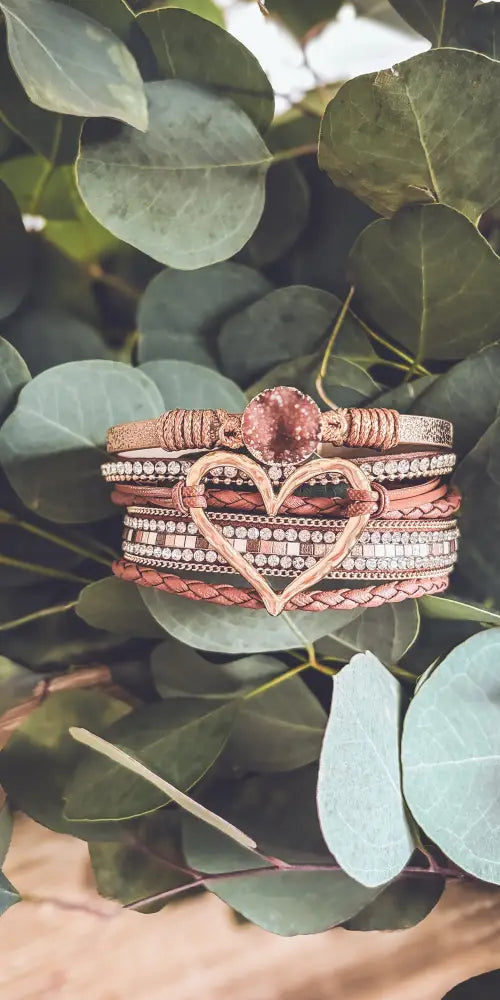 A stack of rose gold braided leather bracelets with a heart charm and sparkling crystal accents