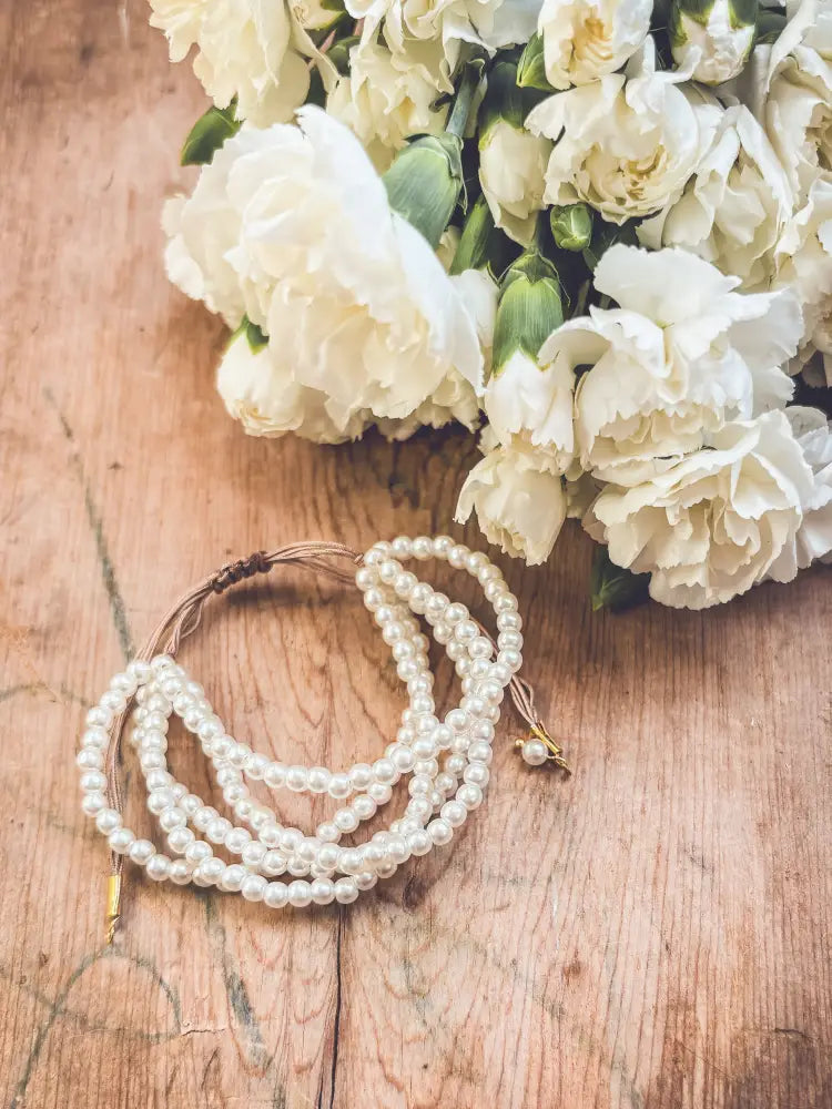 A delicate multi-strand pearl bracelet with braided brown cord and gold clasp resting beside white flowers on rustic wood