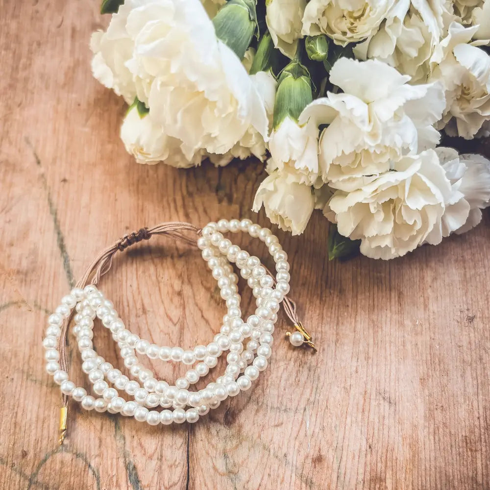Multi-strand pearl bracelet with braided brown cord and gold clasp resting beside white flowers on wooden surface