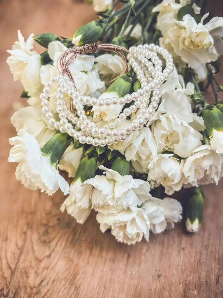 Multi-strand pearl bracelet with braided brown cord and gold clasp resting on white carnations