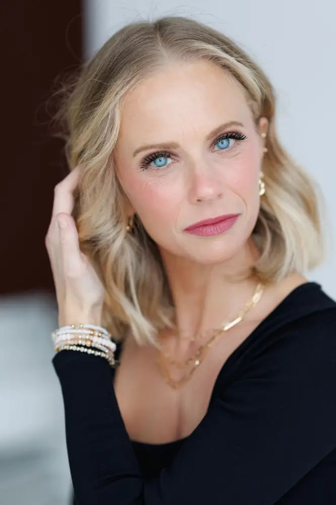 A woman with blonde wavy hair wears an elegant pink and gold beaded stacking bracelet set with delicate gold jewelry