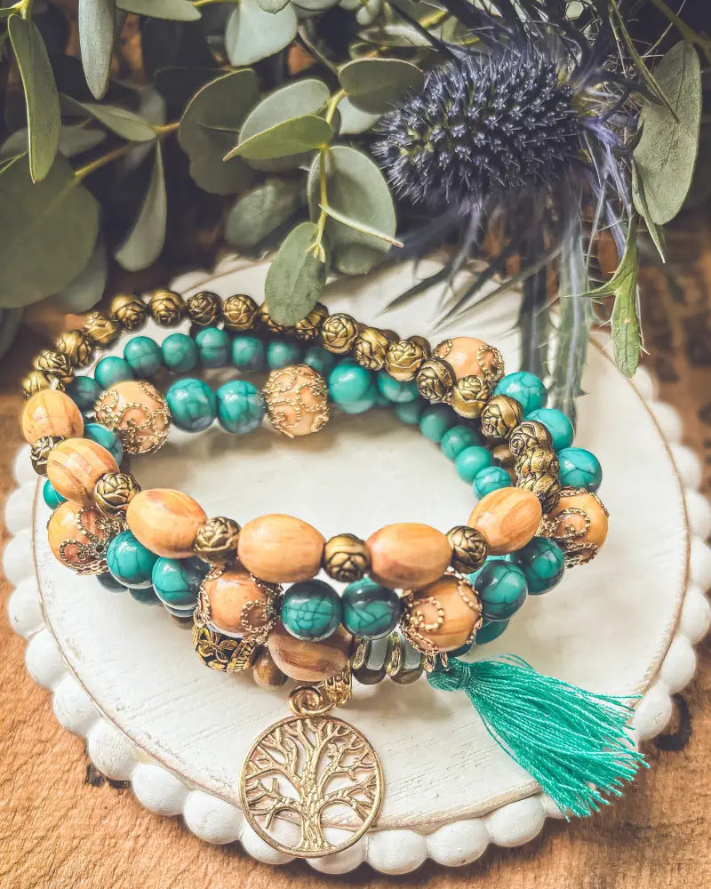A set of three stacked turquoise and warm wood beaded bracelets featuring gold-tone accents and a tree of life pendant