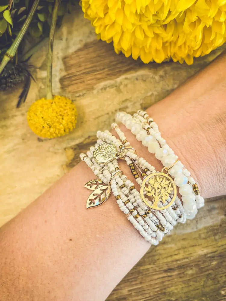 A delicate white beaded wrap bracelet adorned with gold-toned tree of life and leaf charms for stacking