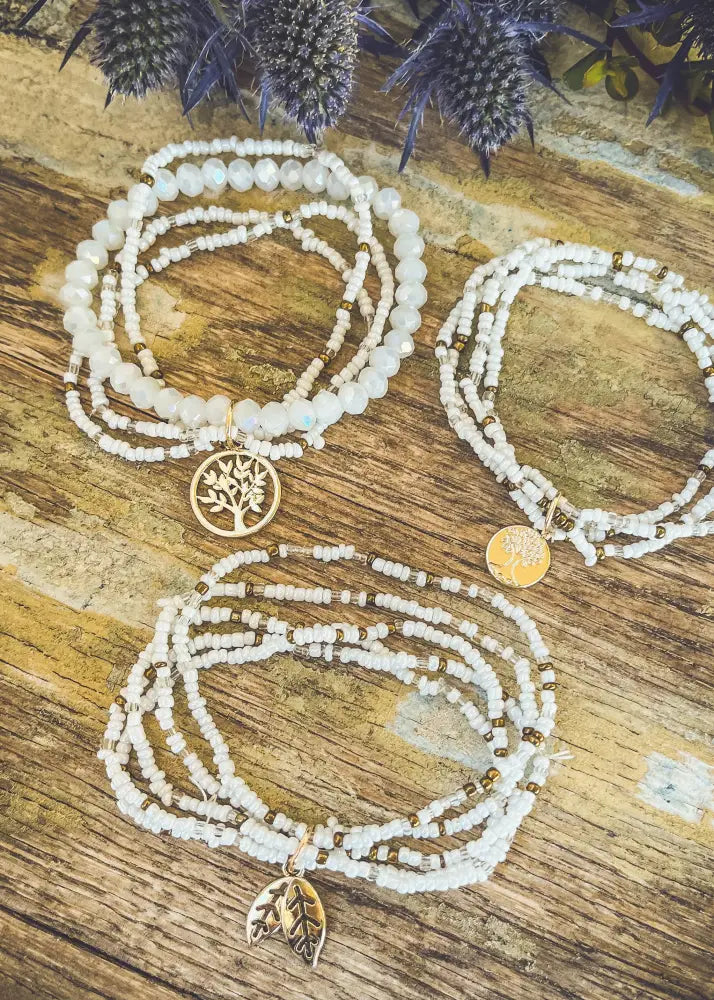 Three layered white and gold beaded stacking bracelets featuring tree of life leaf and circular metallic charms