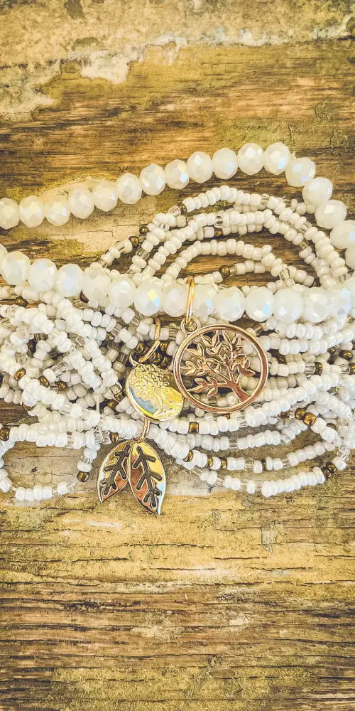 A delicate stack of white beaded bracelets with gold tree of life and leaf charms in a stacking bracelet set