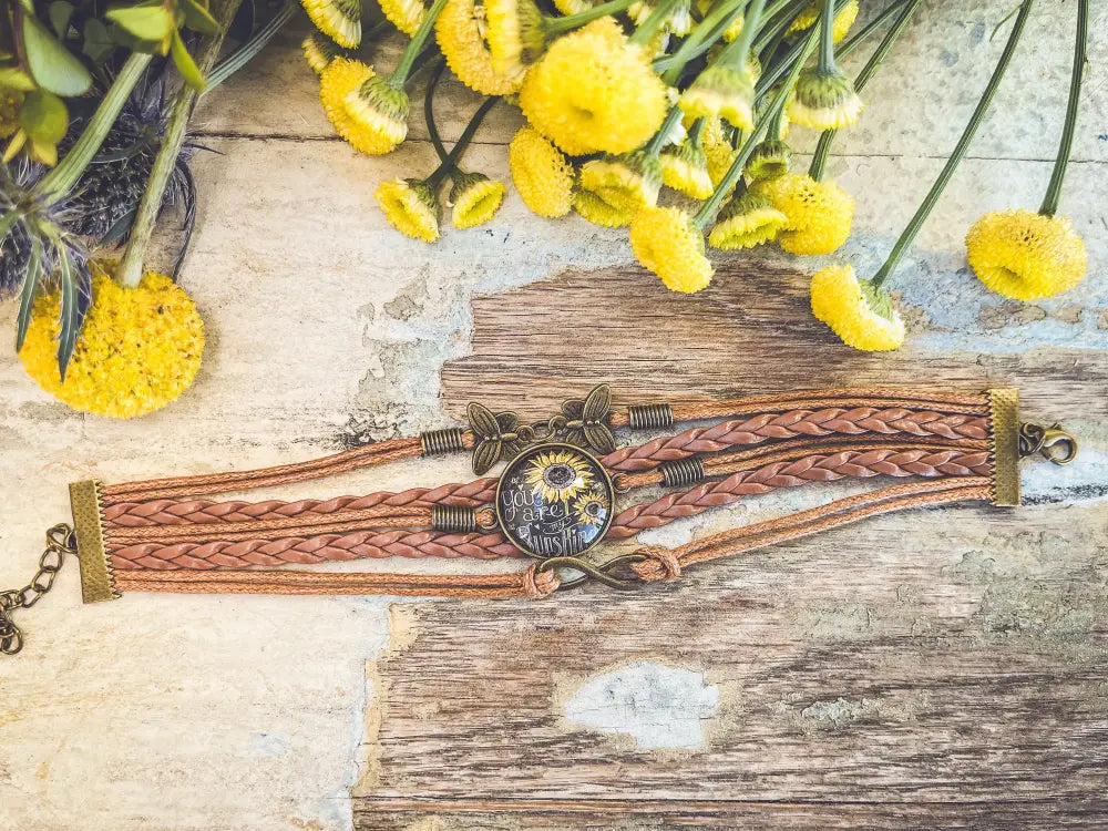 A rustic brown braided leather sunflower bracelet with a glass charm and brass-toned metal accents with an adjustable clasp