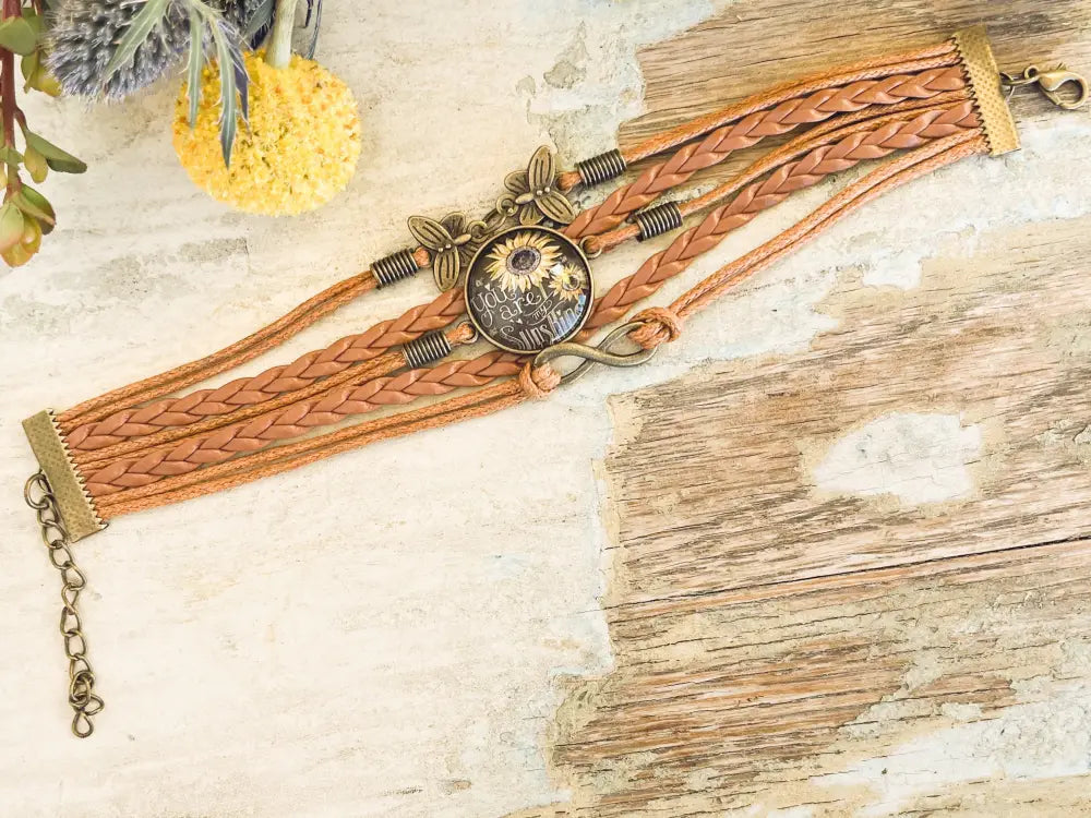 A tan braided leather You are my Sunshine bracelet with a central sunflower glass charm and gold-toned butterfly accents