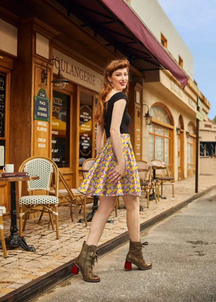 A woman wearing a bees and dandelions pink floral cotton skater skirt paired with a black sleeveless top and olive boots