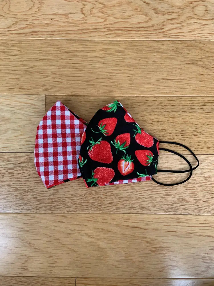 Two fabric face masks reversible with red and white gingham and vibrant red strawberries on black background