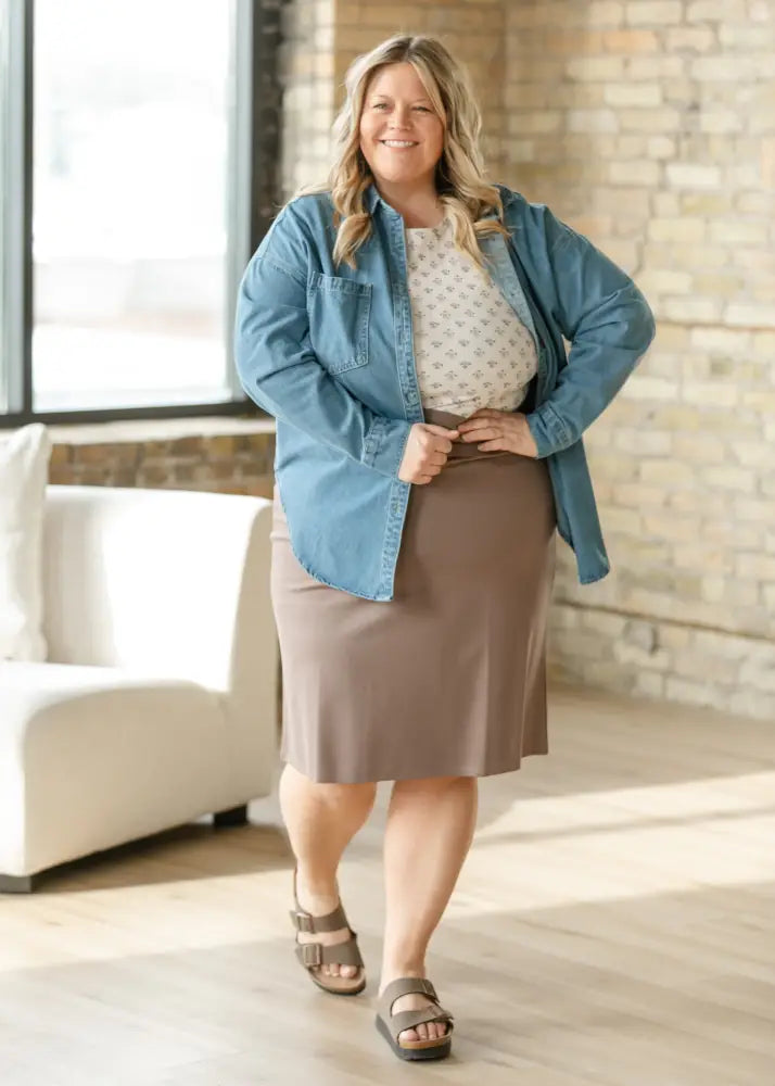A plus-size woman wearing a light blue Betty denim relaxed button up shirt made of denim paired with a taupe skirt