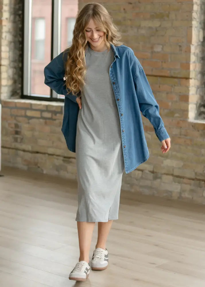 A woman wears the betty denim relaxed button up shirt in oversized blue denim over a light gray knee-length dress