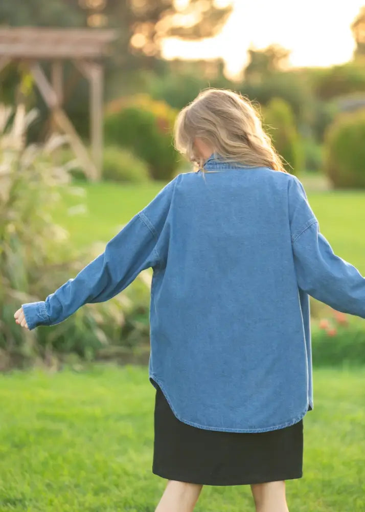 A woman wears a loose-fitting medium-blue denim Betty Denim relaxed button up shirt with long sleeves and curved hemline