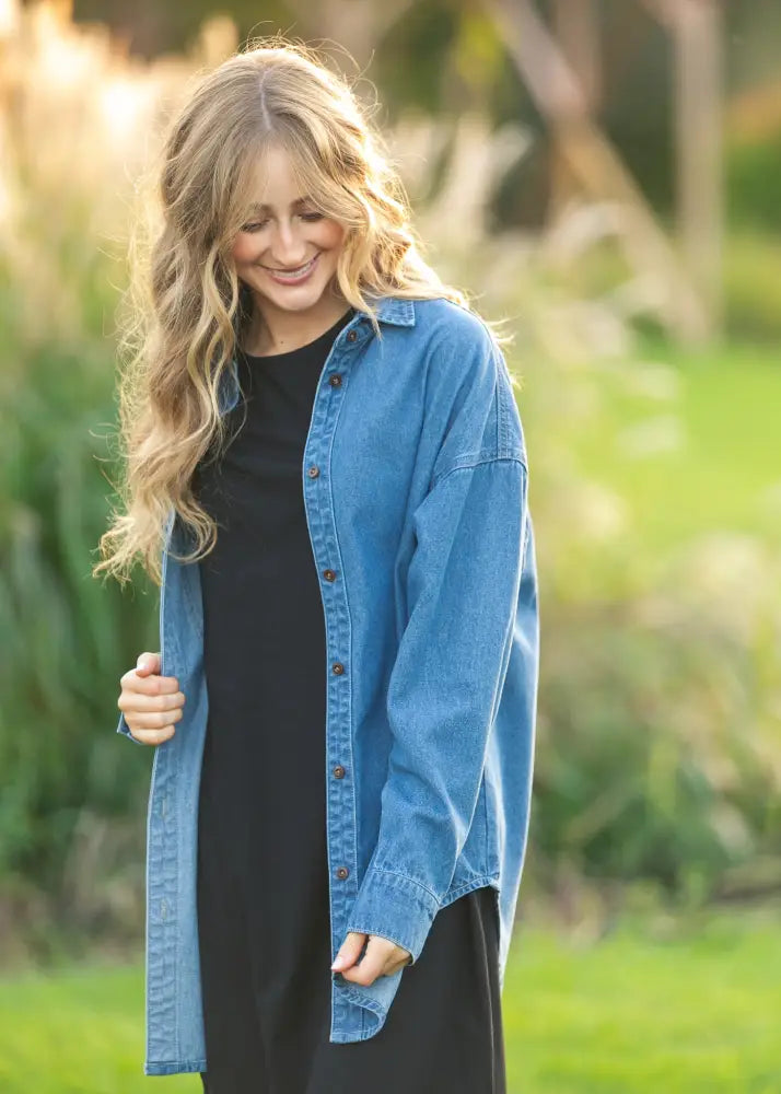 A woman wears a Betty Denim relaxed button up shirt in medium-wash blue denim with button closures over a black dress
