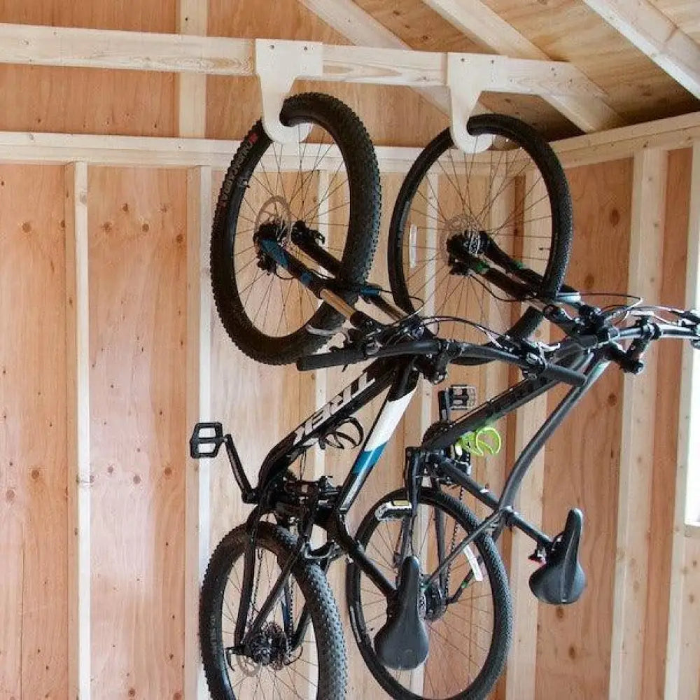 Two black mountain bikes with blue and white accents hang from a wooden shed ceiling on heavy duty vertical bicycle storage hooks