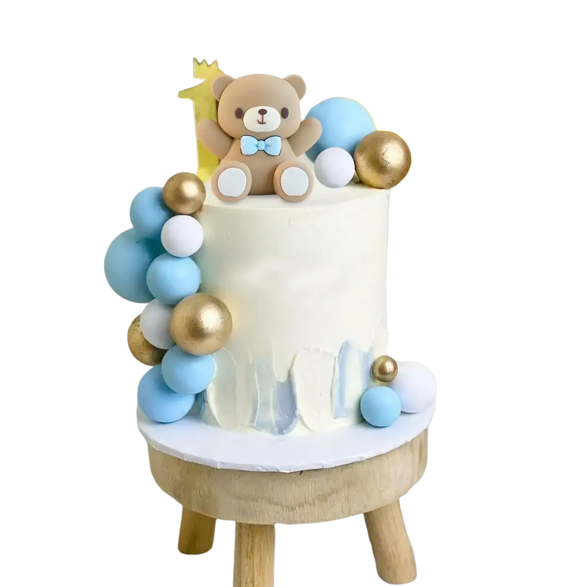 A white frosted cake with a brown teddy bear topper wearing a blue bow tie surrounded by blue gold and white spheres