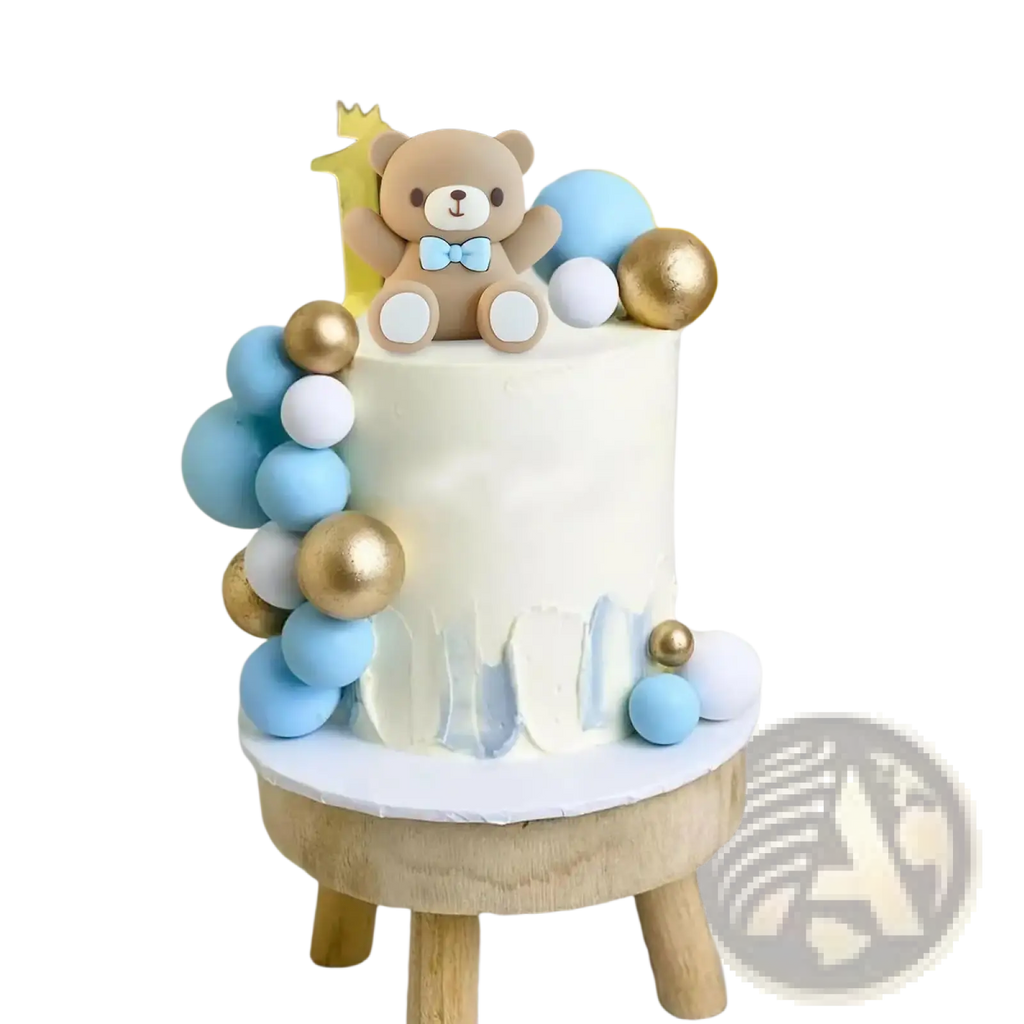 A white frosted cake with a brown teddy bear topper wearing a blue bow tie surrounded by blue gold and white spheres