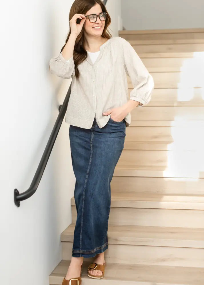 Woman wearing a bria dark wash denim maxi skirt with side slit in dark blue paired with a light beige button-up shirt