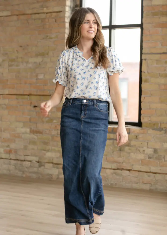 A woman wearing a bria dark wash denim maxi skirt with side slit paired with a white blouse with blue floral patterns