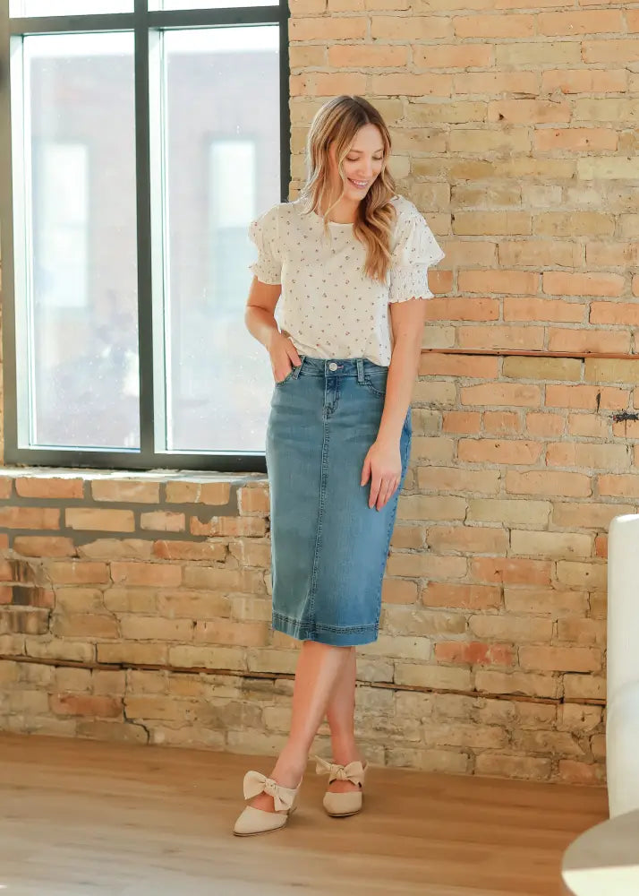 A woman wears a white polka dot blouse with ruffled cuffs and a bryn light wash sustainable denim midi skirt