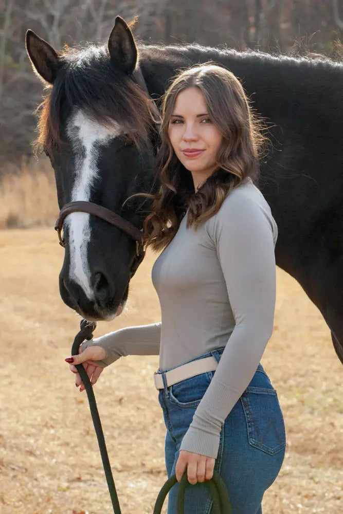 A woman wearing a gray btr active women’s seamless upf 30 sun protection baselayer stands beside a black horse