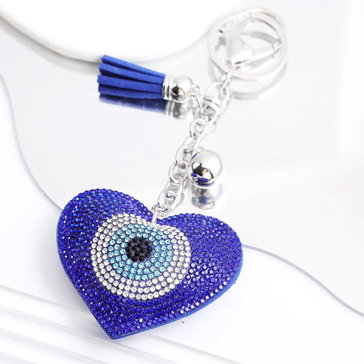 A vibrant blue heart-shaped evil eye keychain with sparkling rhinestones and a leather tassel featuring a detailed concentric circle design in black light blue and white.