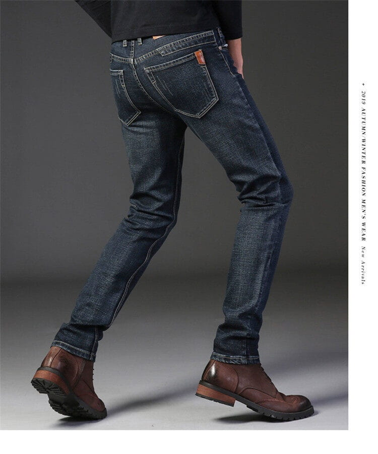 Pantalones vaqueros de mezclilla elástica de corte slim fit para hombre, confeccionados en mezclilla azul marino oscuro, con sutil desvanecido, costuras clásicas y un parche de cuero marrón en el bolsillo trasero, mostrados desde el lado posterior.